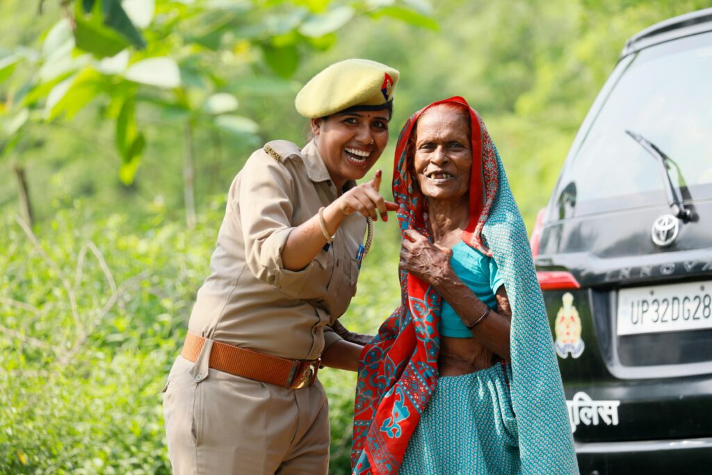 A smiling policewoman helps an elderly woman by a parked police car in a lush setting.